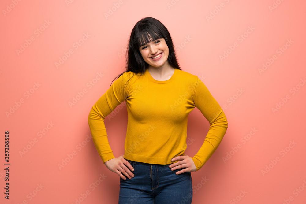 Woman with yellow sweater over pink wall posing with arms at hip and smiling Woman with yellow sweater over pink wall posing with arms at hip and smiling