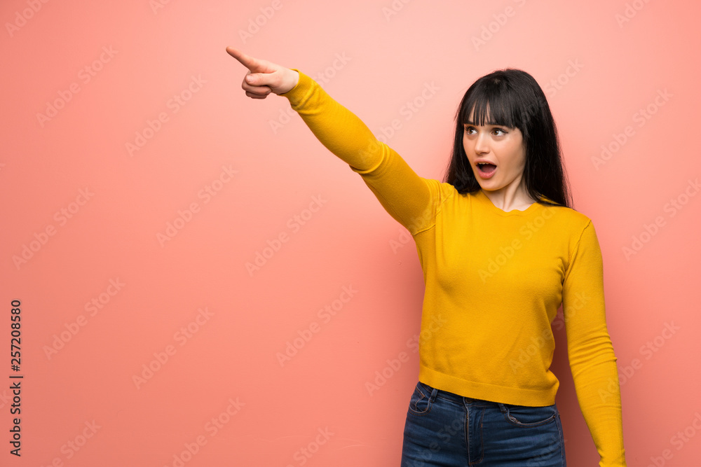 Woman with yellow sweater over pink wall pointing away Woman with yellow sweater over pink wall pointing away