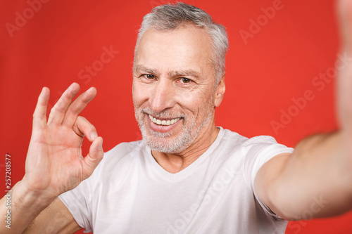 Portrait of happy senior bearded man showing ok sign and taking selfie on smartphone, isolated against red background.