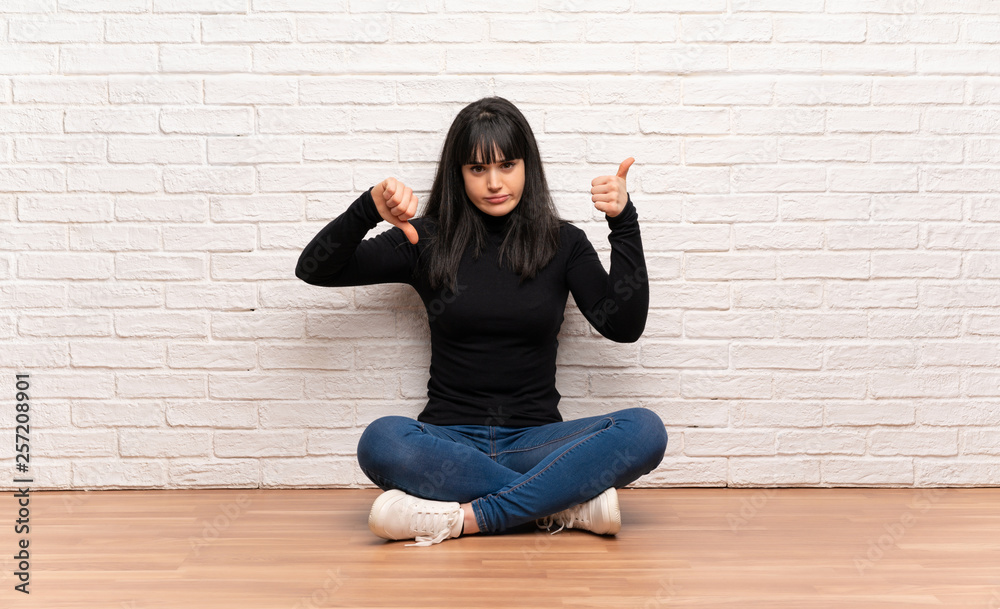 Woman sitting on the floor making good-bad sign. Undecided between yes or not Woman sitting on the floor making good-bad sign. Undecided between yes or not