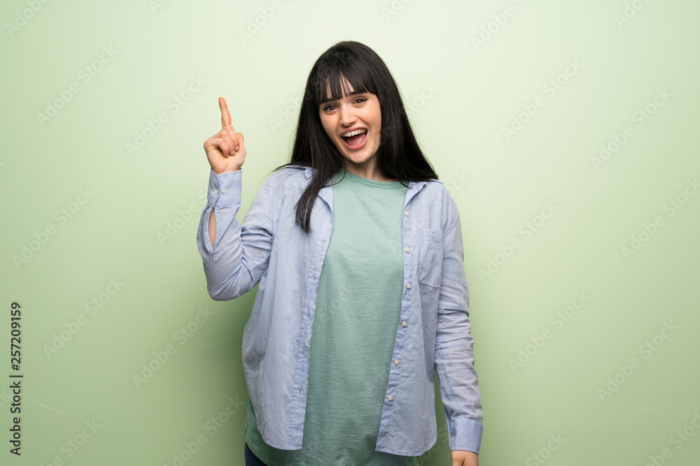 Young woman over green wall intending to realizes the solution while lifting a finger up Young woman over green wall intending to realizes the solution while lifting a finger up