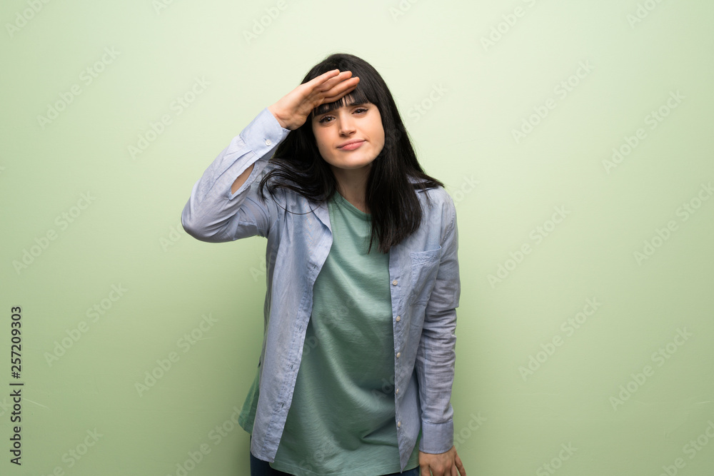 Young woman over green wall looking far away with hand to look something Young woman over green wall looking far away with hand to look something