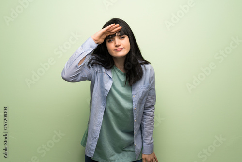 Young woman over green wall looking far away with hand to look something