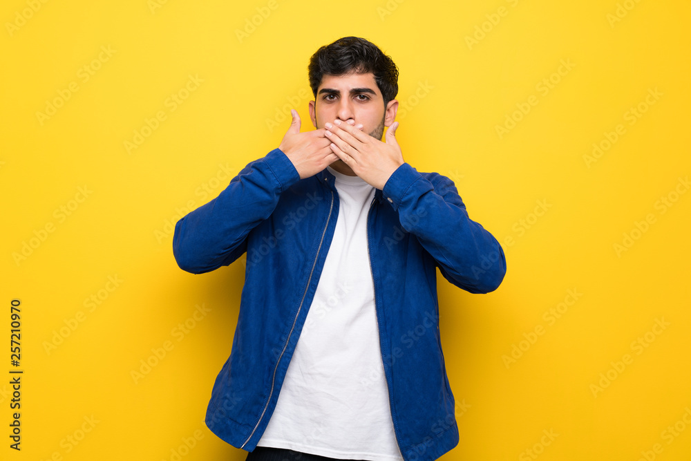 Man with blue jacket over yellow wall covering mouth with hands Man with blue jacket over yellow wall covering mouth with hands