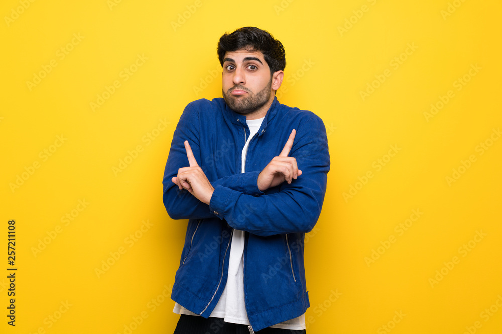 Man with blue jacket over yellow wall pointing to the laterals having doubts Man with blue jacket over yellow wall pointing to the laterals having doubts