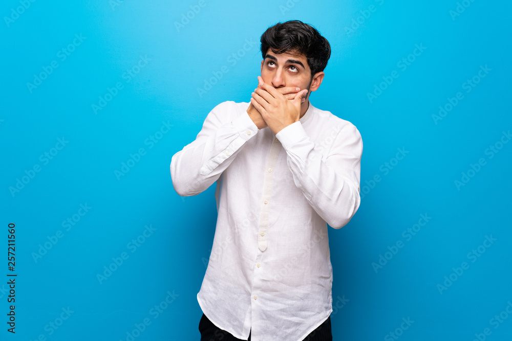 Young man over isolated blue wall covering mouth with hands Young man over isolated blue wall covering mouth with hands