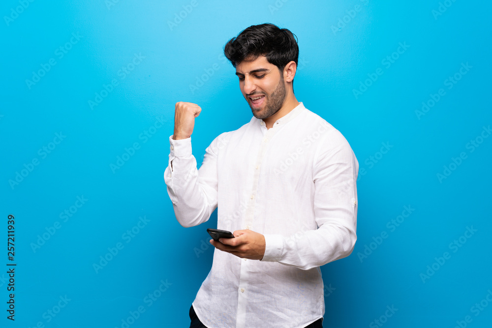 Young man over isolated blue wall celebrating a victory with a mobile Young man over isolated blue wall celebrating a victory with a mobile