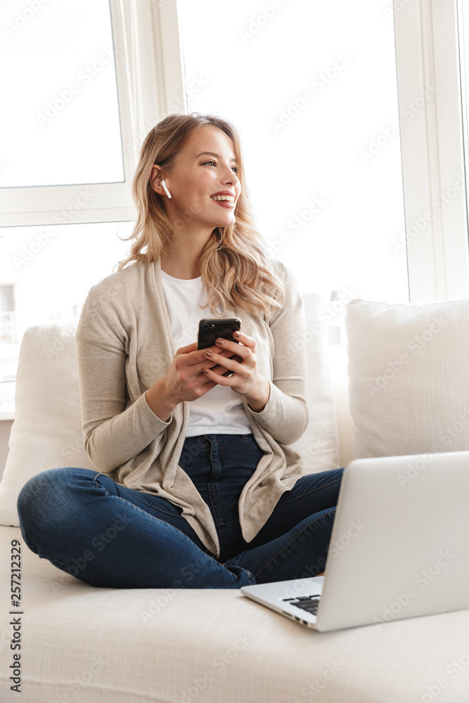 Blonde woman posing sitting indoors at home using laptop computer and mobile phone.