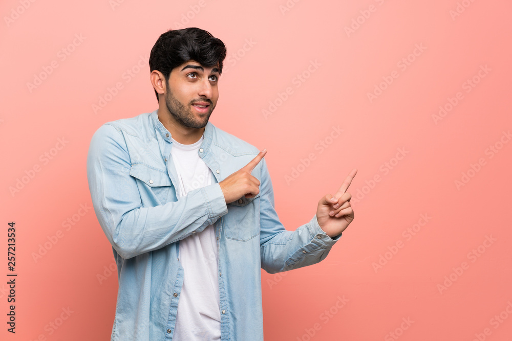 Young man over pink wall frightened and pointing to the side Young man over pink wall frightened and pointing to the side
