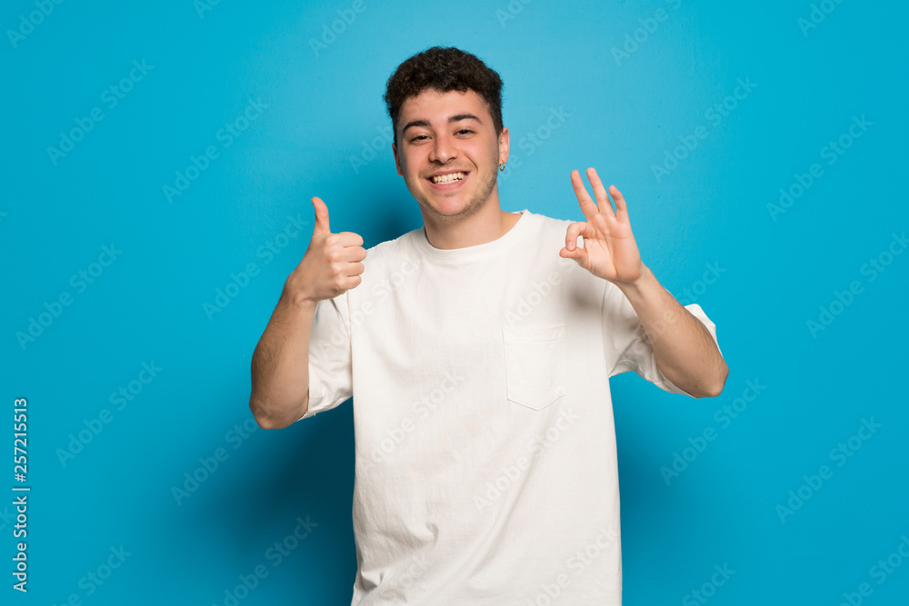 Young man over blue background showing ok sign with and giving a thumb up gesture Young man over blue background showing ok sign with and giving a thumb up gesture
