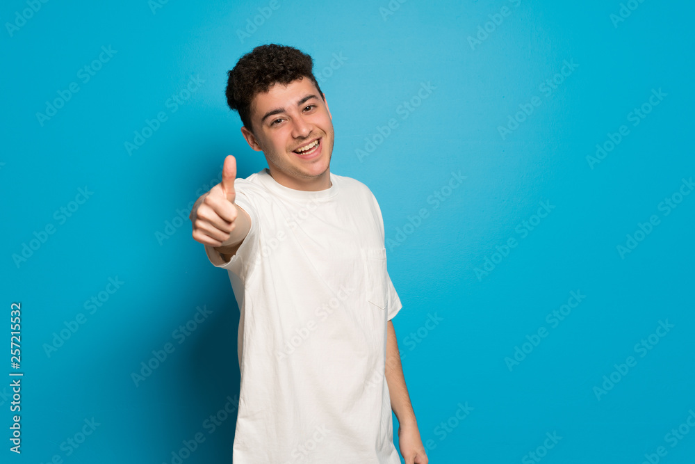 Young man over blue background giving a thumbs up gesture because something good has happened Young man over blue background giving a thumbs up gesture because something good has happened