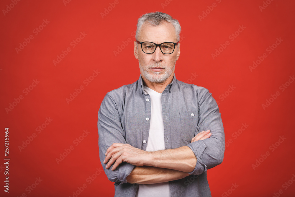 Portrait of a mature serious businessman with crossed hands wearing glasses isolated against red background. Happy senior man looking at camera with copy space. Portrait of a mature serious businessman with crossed hands wearing glasses isolated against red background. Happy senior man looking at camera with copy space.