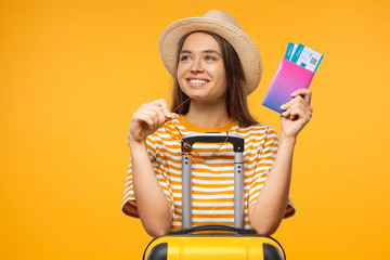 Dreaming young girl holding flight tickets and suitcase, isolated on yellow background. Ready to travel concept