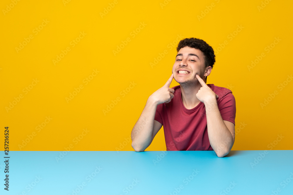 Young man with colorful wall and table smiling with a happy and pleasant expression Young man with colorful wall and table smiling with a happy and pleasant expression