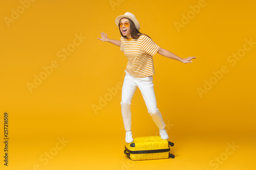 Dream about traveling concept. Young tourist girl standing on suitcase, pretending like she is surfing, isolated on yellow background