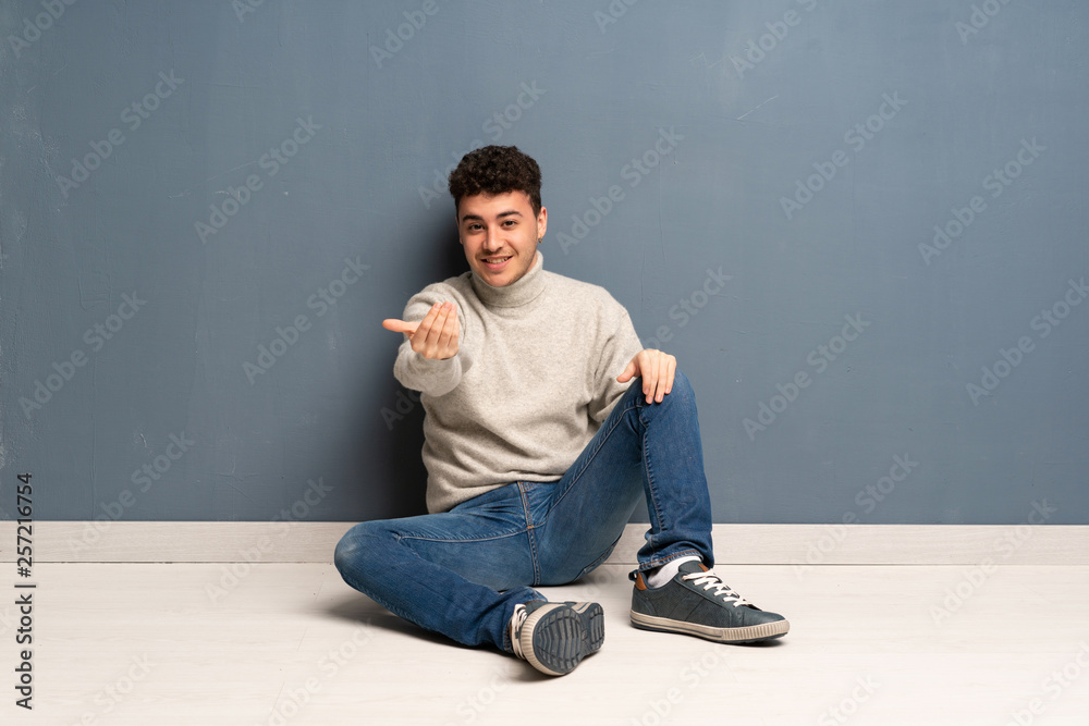 Young man sitting on the floor inviting to come with hand. Happy that you came Young man sitting on the floor inviting to come with hand. Happy that you came