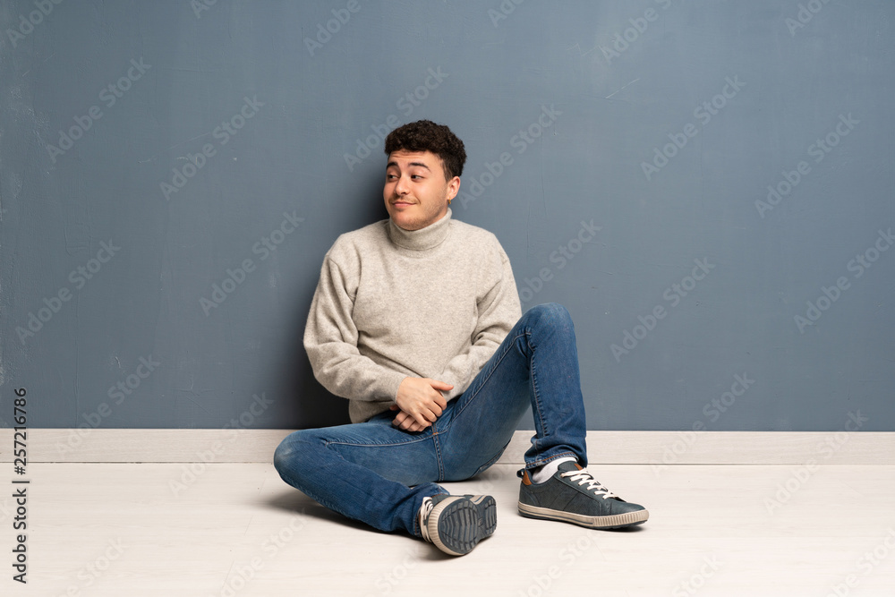 Young man sitting on the floor making doubts gesture looking side Young man sitting on the floor making doubts gesture looking side