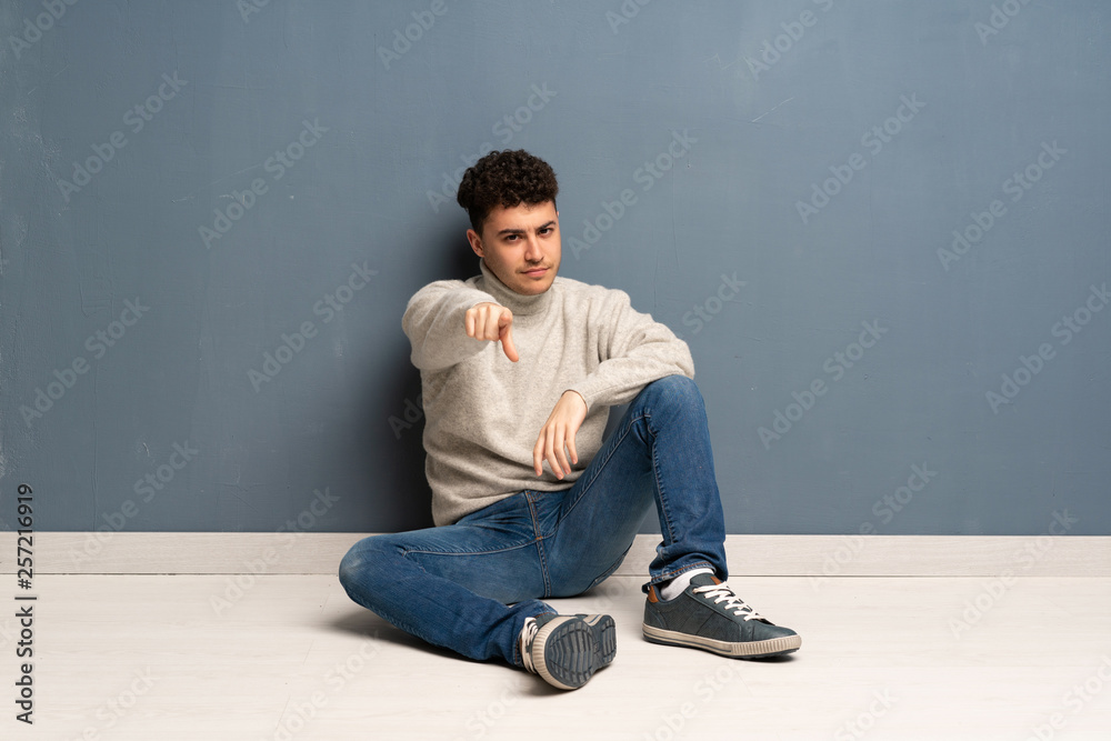 Young man sitting on the floor points finger at you with a confident expression Young man sitting on the floor points finger at you with a confident expression