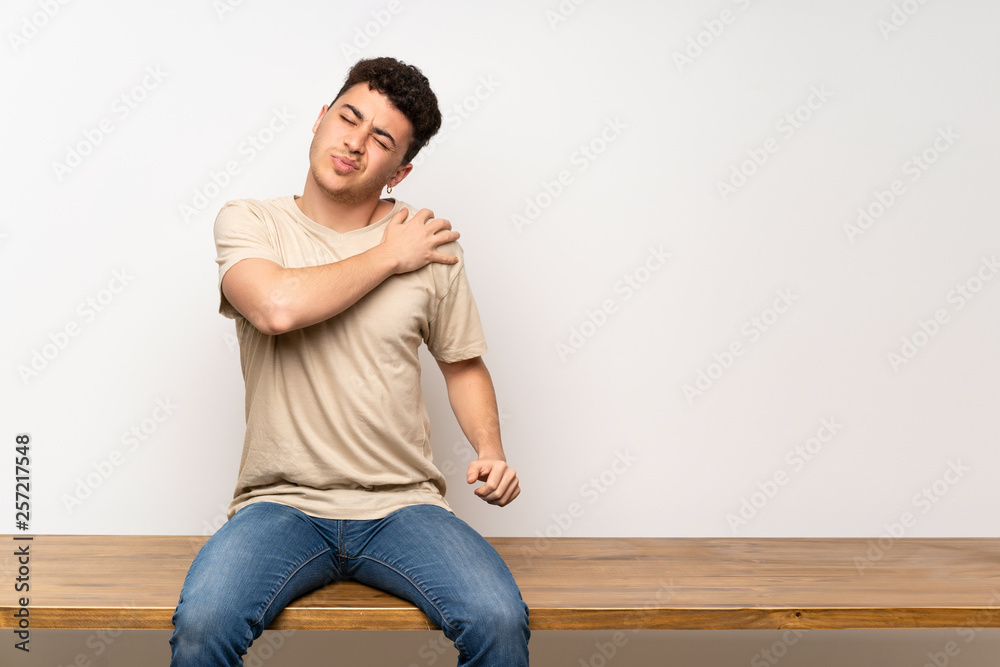 Young man sitting on table suffering from pain in shoulder for having made an effort Young man sitting on table suffering from pain in shoulder for having made an effort