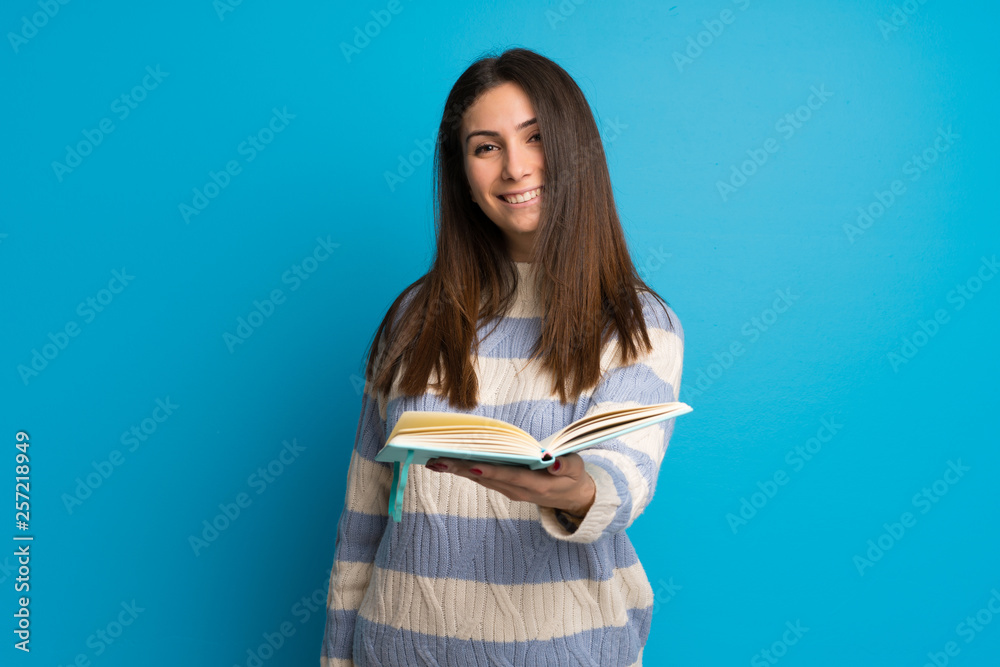 Young woman over blue wall holding a book and giving it to someone