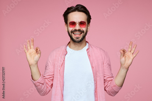 Relaxed young handsome man keeps both hands in okay gesutre, meditates indoor, tries gather his strength, isolated over pink background. Caucasian male in stylish pink shirt and sunglasses gestures. photo