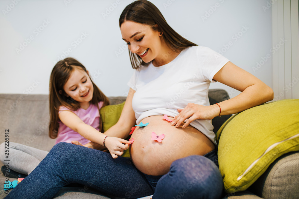 Pregnant woman and her cute daughter putting paper stickers on belly. Concept of choosing baby name. Pregnant woman and her cute daughter putting paper stickers on belly. Concept of choosing baby name.
