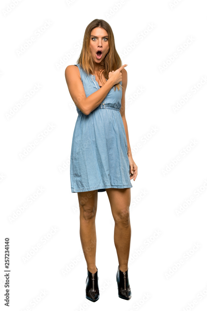 A full-length shot of a Blonde woman with jean dress surprised and pointing side over isolated white background A full-length shot of a Blonde woman with jean dress surprised and pointing side over isolated white background