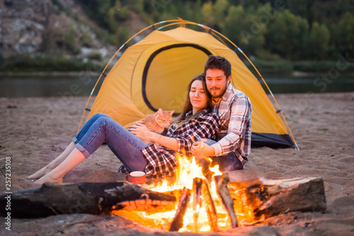 people, summer tourism and nature concept - couple near camp fire warming up seen from the tent