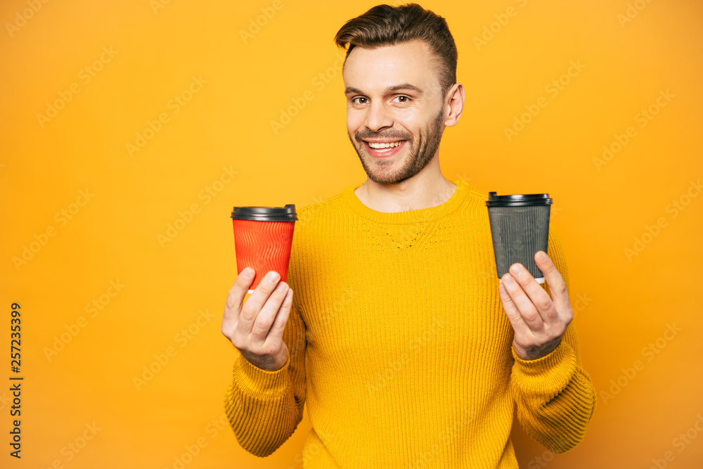 Do you want to have some coffee? Wonderfully looking man in mustard colored sweater is holding two cups of latte in black and red coffee cups in front of rich yellow background. Do you want to have some coffee? Wonderfully looking man in mustard colored sweater is holding two cups of latte in black and red coffee cups in front of rich yellow background.