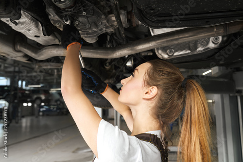 Girl working undercarriage of automobile, using tool.