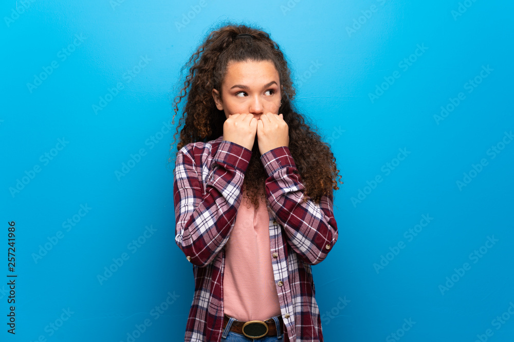 Teenager girl over blue wall is a little bit nervous and scared putting hands to mouth Teenager girl over blue wall is a little bit nervous and scared putting hands to mouth