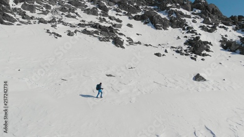 Aerial view of young man hiking climbing snowy mountain at beautiful winter day. Happy active lifestyle video. Male mountaineer with trekking poles and a backpack walking on mountain ridge.