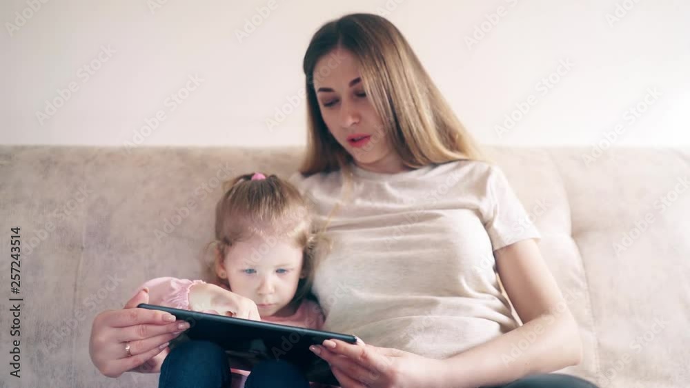 CU, 4k: Young attractive mother and sweet daughter are sitting on the couch and training using a tablet computer. The daughter thinks and taps. CU, 4k: Young attractive mother and sweet daughter are sitting on the couch and training using a tablet computer. The daughter thinks and taps.