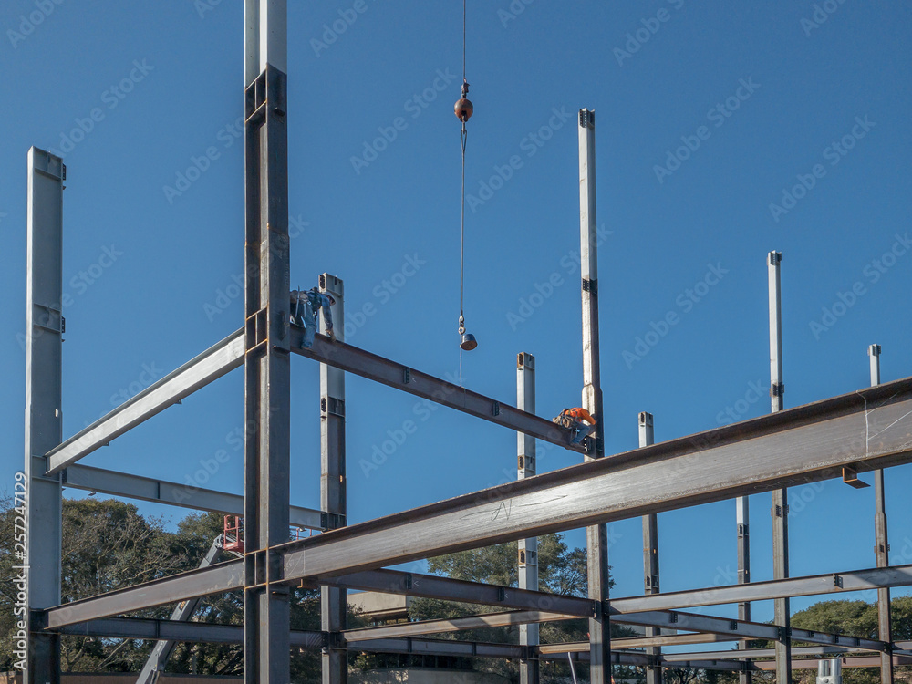 2 ironworkes are on a horizontal steel I-beam just put into place. One is putting in bolts into the beam. The other is about to crawl to the cable. Work on the steel structure of a building. Blue sky. 2 ironworkes are on a horizontal steel I-beam just put into place. One is putting in bolts into the beam. The other is about to crawl to the cable. Work on the steel structure of a building. Blue sky.