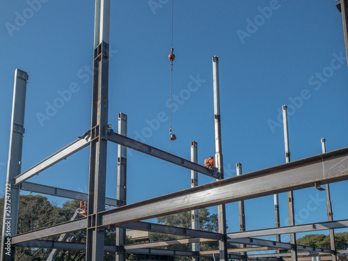 2 ironworkers are on a horizontal steel I-beam just put into place. Both are beginning to put bolts at each end of the beam. 2 working on the steel structure of a building. Blue sky is behind.