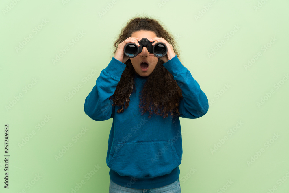 Teenager girl over green wall and looking in the distance with binoculars Teenager girl over green wall and looking in the distance with binoculars