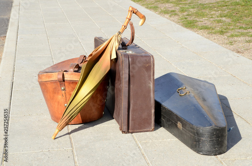 Photography Vintage luggage items, suitcase, umbrella, hat box and musical instrument standing on a sidewalk in sunlight