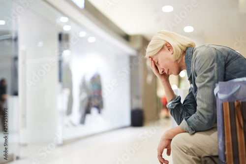 Serious exhausted mature lady tired from shopping sitting on bench in mall and leaning head on hand while keeping eyes closed, she feeling headache