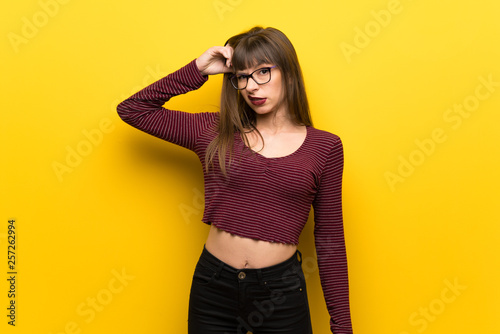 Woman with glasses over yellow wall having doubts while scratching head