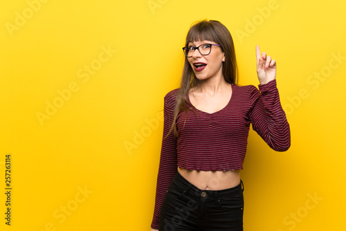 Woman with glasses over yellow wall intending to realizes the solution while lifting a finger up