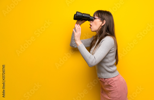 Woman with glasses over yellow wall and looking in the distance with binoculars © luismolinero