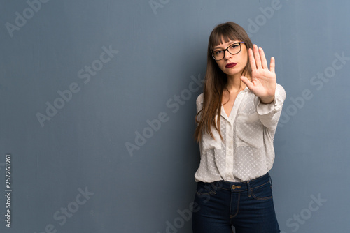 Woman with glasses over blue wall making stop gesture denying a situation that thinks wrong