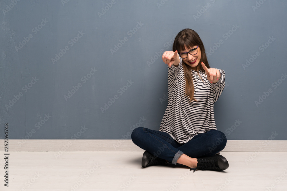 Woman with glasses sitting on the floor points finger at you while smiling Woman with glasses sitting on the floor points finger at you while smiling
