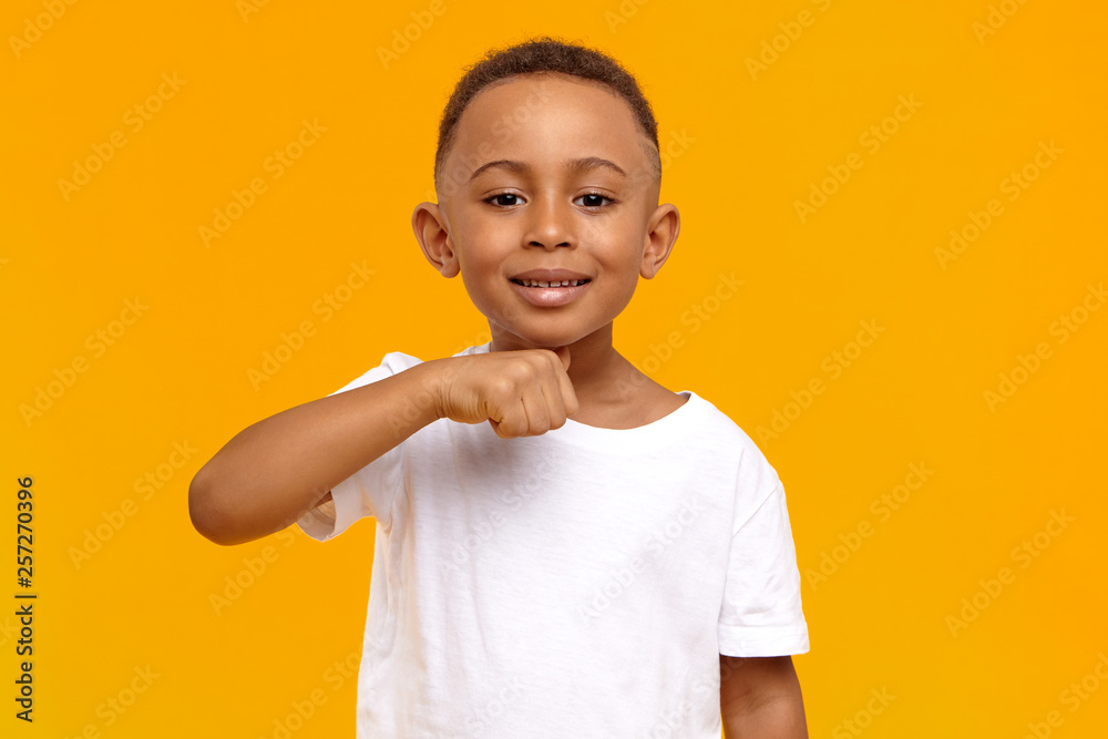 I will cut your head off. Picture of positive cheerful dark skinned schoolboy having fun in yellow studio, making joke, holding thumb at his neck, showing menacing warning gesture, smiling broadly - obrazy, fototapety, plakaty 