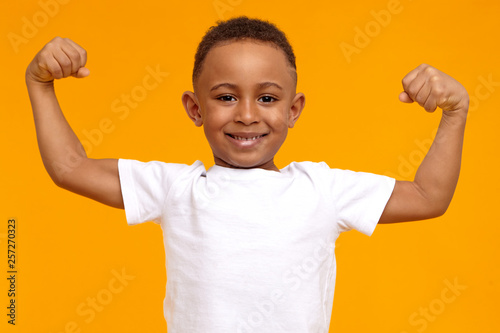 Handsome confident Afro American eight year old child in casual t-shirt smiling happily and raising clenched fists, tensing muscles, feeling strong and full of energy after ate healthy protein lunch
