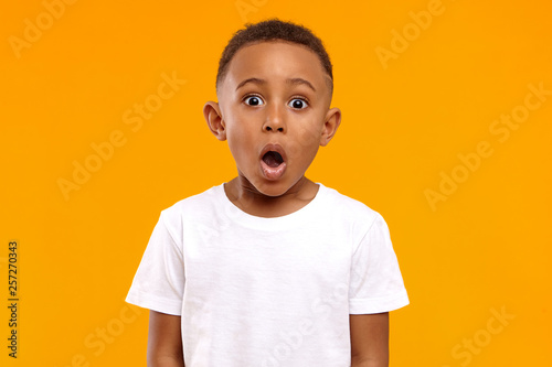Surprise, excitement and fascination concept. Funny bug eyed African little boy opening his mouth widely, shocked with astonishing unexpected news, having amazed look, showing full disbelief