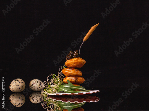 Easter delicate snack with black caviar, quail eggs, poffertjes, alfalfa, rucola over black background