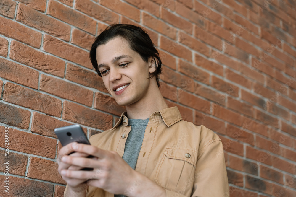 © Oleksii - Happy young man using mobile phone and internet,  streaming video online, reading news, watching movie. Stylish hipster in casual shirt holding smartphone, chatting, laughing, standing near brown wall