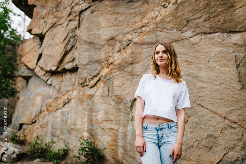 Slim girl stands and poses at cliff. Portrait of a woman in jeans and a white T-shirt, against the background of the mountain on nature. upper half. Looking sideways. Close up
