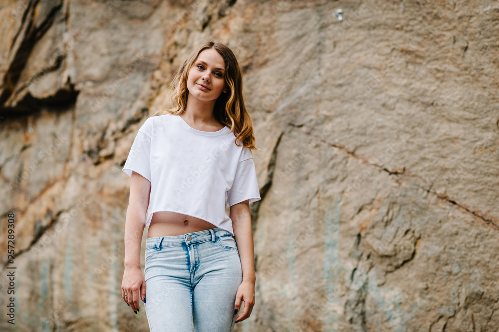 Slim girl stands and poses at cliff. Portrait of a woman in jeans and a white T-shirt, against the background of the mountain on nature. upper half. Looking at the camera. Close up